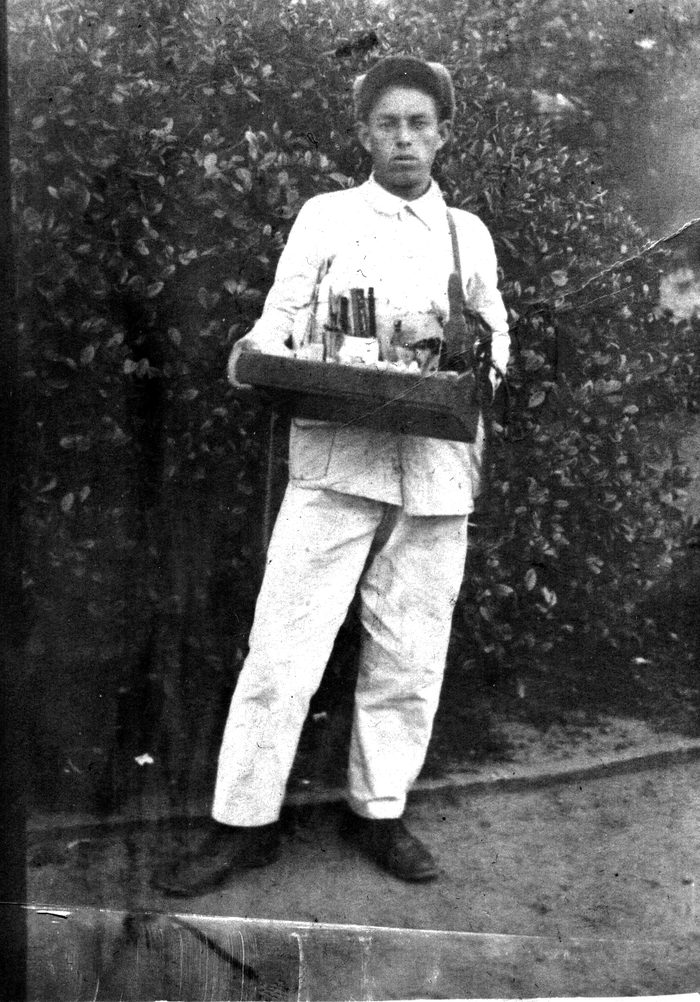 Un joven vestido de blanco, con gorra y una bandeja de mercadería colgada del hombro, de pie frente a un seto.